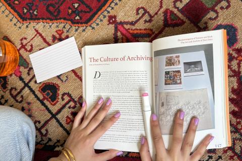 The Great Book Return. Open face book reading The Culture of Archiving with picture on right hand side, being held open by hands with light pink nails holding pink highlighter wearing gold bracelet on left hand. Blank lined cue cards lay next to the book, with background red, white rug with intricate lines and shapes. 
