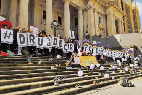 A crowd of people gather on the steps of Parliament House holding signs with individual letters that spell out "# DRUG DECRIM TO END OVERDOSE".
