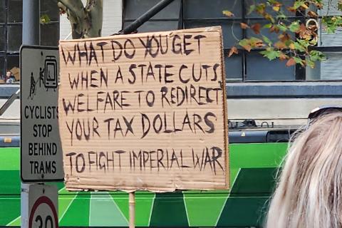 Photograph of a handwritten carboard protest sign at the NDIS rally in Naarm 2026 reading "WHAT DO YOU GET WHEN A STATE CUTS WELFARE TO REDIRECT YOUR TAX DOLLARS TO FIGHT IMPERIAL WAR".