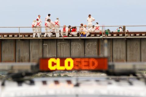 A photograph of protesters dressed in white jumpsuits with red 'x' on the back blocking a railway bridge in Newcastle. A sign on the side of the bridge reads 'CLOSED'.