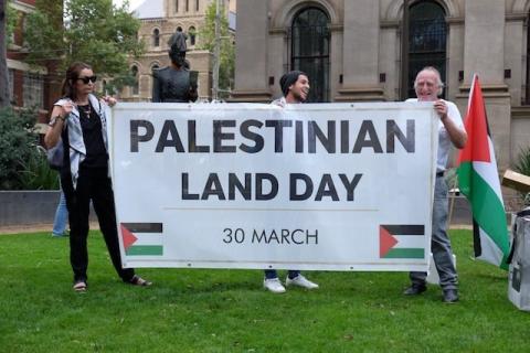 A Palestininan Land Day banner being held by three people. The banner reads "Palestininan Land Day 30 March" and is decorated with Palestinian flags.