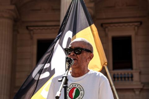 Professor Gary Foley speaking at the Naarm Invasion Day Rally 2026. He is wearing a shirt with a map of Palestine and the Camp Sovereignty flag flying behind him.