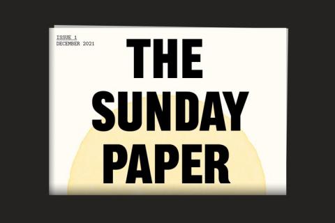 A digital image of The Sunday Paper, a folded newspaper on a black background. The cover has 'The Sunday Paper' written across it in large black capital letters, with half a yellow painted sun rising from the fold behind the text. In the top left corner is printed 'Issue 1 December 2021'.