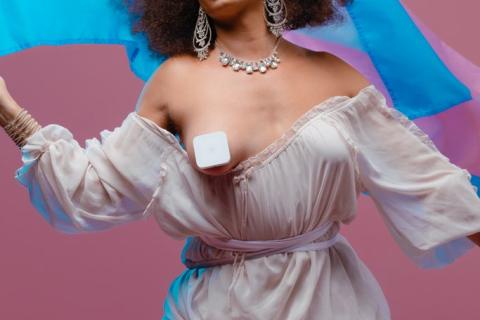 A photograph of Miss Cairo wearing a ruffled, flowy pale pink dress pulled down in front to reveal one censored breast. She holds the trans flag up with one hand, and it waves behind her head. She is wearing elaborate crystal earrings, a necklace and a bracelet.