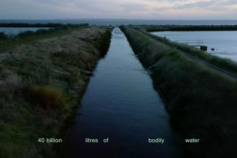 A cinematic image of the canals and grassy edges of the Western Treatment Plant on Wadawurrung Country, with text that reads "40 billion litres of bodily water". Image Credit: Eugenia Lim, Metabolism, 2023, 2K video, colour, sound, 29:15. Cinematographer: Tim Hillier.