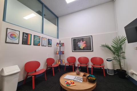 A photograph of the CanTEST Health and Drug Checking service waiting room. The room has red chairs lining the wall, a low table, and posters on the wall with health and harm minimisation information about drugs.