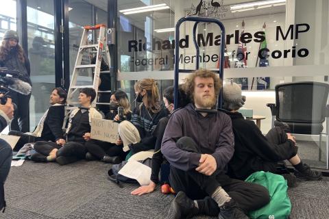 A group of protesters sit on the floor outside Richard Marles' office. Some of them are locked on by the neck to a trolley, while others are locked on by the neck to a ladder standing in the open doorway of the office.