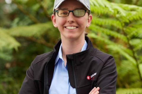 Cassie Hames is wearing a cap and black jacket saying Sage Automation with her arms folded with a content smile on her face standing in front of Green plants.