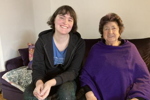 Amy and Aunty Lyn sitting next to each other on a couch smilling for a photo graph. Amy has an opened, black zipper hoodie with a 3CR T-Shirt. She has brown hair in a bob with bangs. Aunty Lyn is smiling away in a purple poncho with short dark hair with light purple hair dye. 