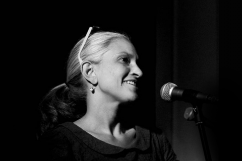 Black and white photograph of a woman speaking into a microphone. Only her face is clearly visible. She has fair hair and slender features.