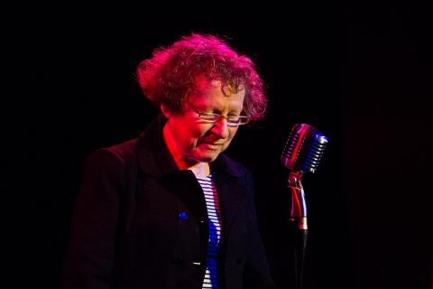 Picture of Ania Walwicz, an older person with olive skin and light hair, resting on stage in front of a microphone. She is about to speak.