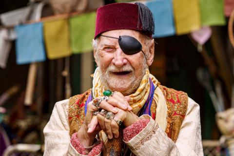 A bearded elderly man wearing a burgandy velvet fez and an eye patch. He has many rings on his fingers, is holding a staff and is wearing brightly coloured clothing. He is also smiling.