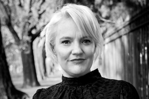 Jacinta Parsons is a caucasian woman with short platinum blonde hair, she wears a black lacy short sleeved top and black trousers and dark lipstick, the photo is black and white, in the image she is standing outside in a park with trees behind her as she smiles at the camera.