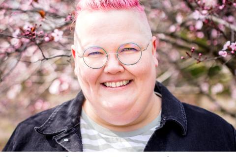 Jasper Peach is a Non-Binary Trans caucasian person with short pink spiky hair, wearing a grey shirt with a dark collared shirt over the top, they stand smiling against a cherry blossom tree outside.