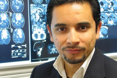 A man with dark hair stands in front of some x-rays of brains. Brainwaves Logo in blue and purple is below the image in a banner.