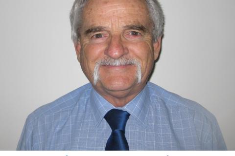 Neil Hunter is a caucasian man with grey hair and a moustache, wearing a powder blue collared shirt and a navy blue tie, he smiles at the camera against a white wall.