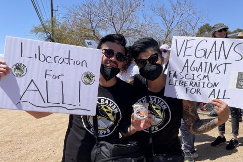 Photo of Claire and Jesse smiling and holding up signs saying Liberation for All and Vegans Against Fascism