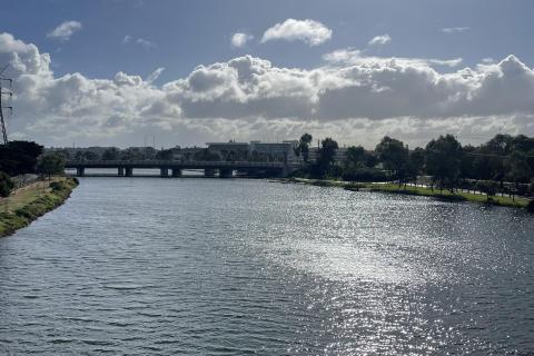 A photograph of the Maribyrnong River, on a sunny and partly cloudy day, with the sun reflecting on its surface, a bridge in the distance and trees on either side.