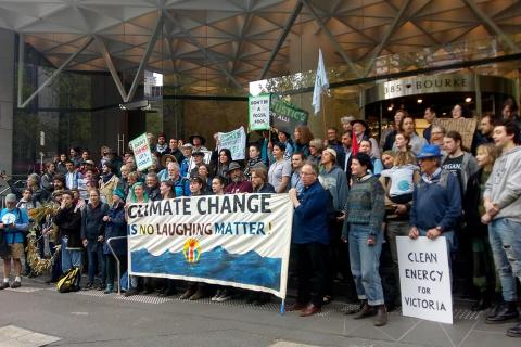 A large group of activists in front of a building holding a "Climate Change is No Laughing Matter" banner