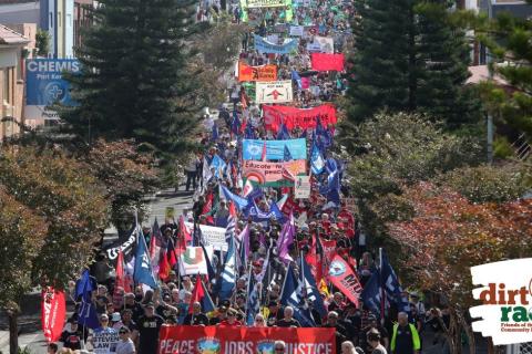 Large crowd with many banners walking through the tree lined streets of Port Kembla main street