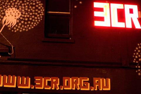 An image of 3CR's building at night time with a mural of a dandelion on the side and large neon sign which reads 3CR, with red text digitally added which reads www.3cr.org.au