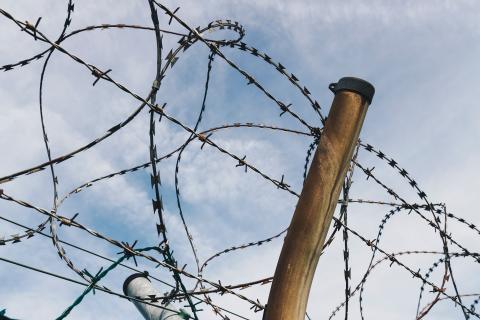 A close-up photograph of a chain-link fence with loops of barbed wire with the sky visible in the background