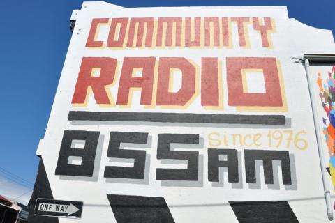 A photograph of the side of 3CR building on a sunny day with a text-based mural on the side which reads: Community Radio, 855AM