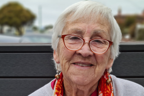 Close-up portrait of the artist. She is an older woman with short silver hair wearing glasses and dangling earings, a colourful orange scarf with a pale blue sweater, and smiling.