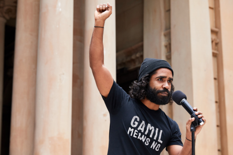 Upper torso & headshot of rally speaker Gamilaraay activist Ian Brown wearing a black t-shirt that says “Gamil means No” and holding microphone while raising fist defiantly. His facial features are serious, sad and grim. The background framing (soft focus) is a section of tall concrete columns and archways at the front of the Sydney town hall.