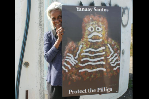 A slender but very strong looking mature Gomeroi Woman is standing in front of a wall that appears to be part of some kind of holding tank. She is holding a sign that says “Yanaay Santos, Protect the Pilliga” and an image invoking a traditional style depiction of the face and shoulders of a humanoid figure using cultural designs. The woman is wearing loose casual clothing (black pants and blue shirt); her hair is silver white and tied back, and she is smiling.