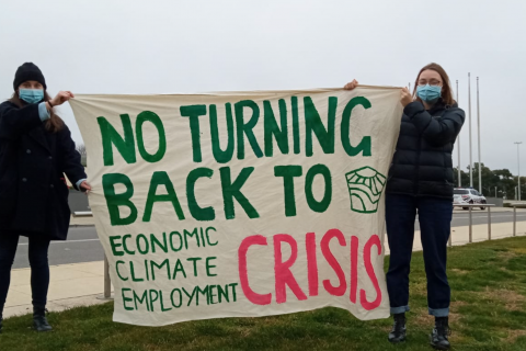Two young people wearing dark toned warm winter clothes and covid protection face masks are holding a banner up on the side of a busy highway. The banner says “No Turning Back to economic, climate, employment CRISIS”.