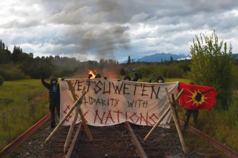 Several anonymous masked protesters wearing black pants, black hoodies, gloves and dark sunglasses stand defiantly with fists in the air at a barricade that has been erected across train tracks in a remote area. One of the protesters is holding up a Wet'suwet'en flag, which has a red background with a yellow sun, and an image of the profile of a Wet'suwet'en warrior wearing headdress in the centre of the sun. The barricade is constructed with 2 large, solid wooden tripods across tracks and a large banner st