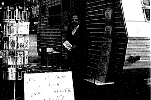 Dark, badly photocopied image showing a man handing out pamphlets outside a caravan in the street.
