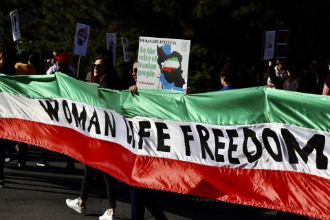 Protesters hold a banner in the colours of the Iranian flag that stretches across the picture with the words 'Women, Life, freedom', in the background people are marching with placards.