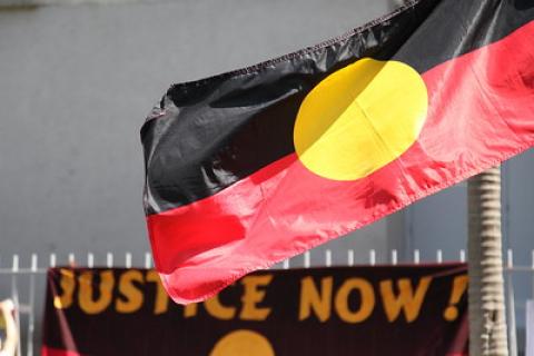 Photo of an Aboriginal flag with the top of protest sign in the background that reads "Justice Now"
