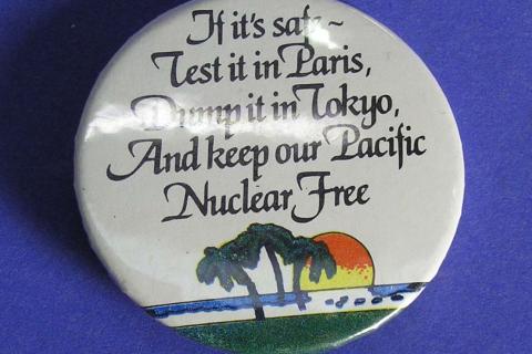  Anti-nuclear protest button badge; black lettering on white ground, "If it's safe - - Test it in Paris - Dump it in Tokyo - And keep our Pacific - Nuclear Free" above Pacific island scene of sun setting behind coconut palms