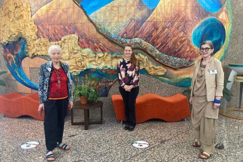 Three women, standing spaced out in front of a mural of mountains.
