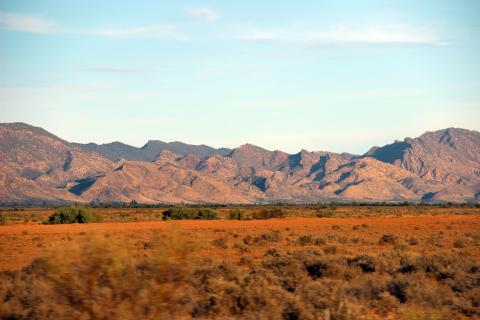 Image taken from moving vehicle, so dry red earth and grasslands in foreground are slightly blurred. Behind are dramatic hills with many peaks, bush covered at the back and less bushy, and dry looking in front.