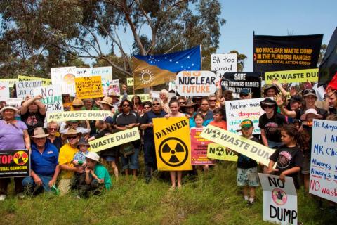 A large crowd of people of all ages outside, in front of a gum tree, holding protest signs that read "No dump", "Danger", "No Cultural genocide", "No nuclear future", "It's Adnyamathanha land Mr Chapman - always was, always will be - No waste dump". There is a Adnyamathanha flag being held up in the centre and an Aboriginal flag held on the right.