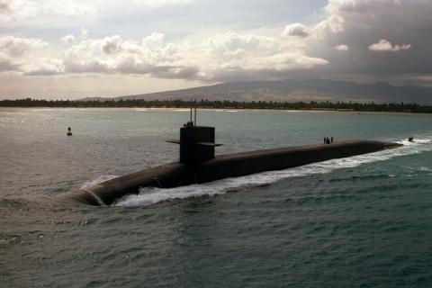 Image of large black nuclear submarine in the centre of the photograph, travelling on the surface of sea. There is a forested coast line in the background and a mountain can be seen in the distance as well as a cloudy sky above.