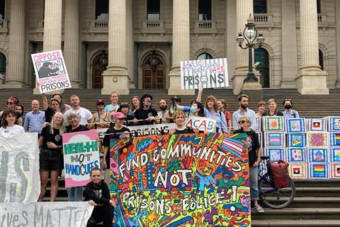 People gathering on the steps of Parliament in Naarm Melbourne with colourful banners says Homes Not Prisons or Police 