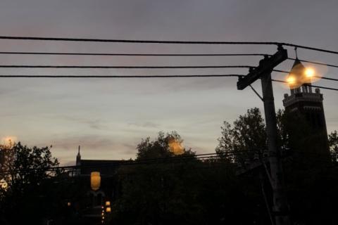 A silhouetted urban street at dusk. The tops of buildings, trees and powerlines can be seen against a grey evening sky.