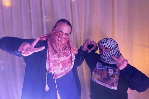 Hosts (left-to-right) Vittorio and Karina wearing Red and Black embroidered Kufiyas, Palestinian Scarves, in front of a semi-transparent curtain, making a love heart out of their respective left and right hands. Vittorio is making a peace symbol with his free hand, while Karina puts up the rude middle finger.