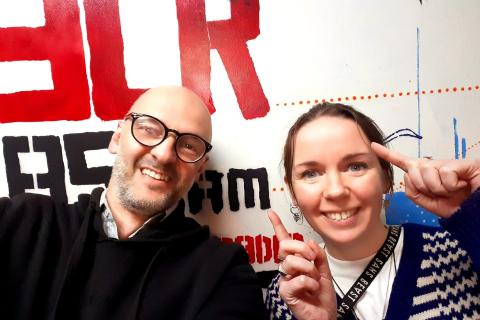 Photograph of presenters Brendan Bonsack (Left) and Carmen Main (Right), smiling at the camera. Behind them is a partial wall mural with the 3CR Radio logo.