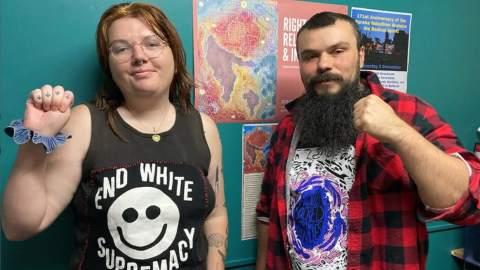 Image description: Em Randall, L, with light-coloured hair, wearing a t shirt that says ‘End White Supremacy’, standing next to Keiran Stewart-Assheton, R. They are in the 3CR studio with fists proudly raised in front of the disability day artwork by Renay Barker-Mulholland.