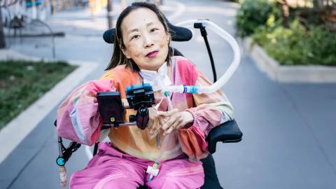 Image of Alice Wong, wearing a pink jumpsuit. She is sitting in a wheelchair with her mechanical ventilator. She is smiling at the camera. 