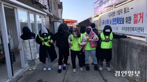 Image of five protesters locking arms in Paju's red light district in South Korea. They are wearing yellow neon vests and covering their face.