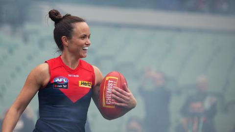 A close up photograph of Melbourne Demons AFLW captain Daisy Pearce standing in a stadium and holding a football in her left arm. She is smiling and wearing a Demons guernsey and her mouthguard. The background is hazy with some spectators visible.