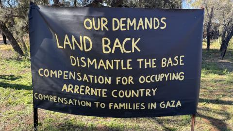 A photo depicting a banner with a list of demands from traditional owners on Arrente Country. There demands are listed as Land Back, dismantly the base, compensation for occupyin Arrernte Country and compansation to families in Gaza.