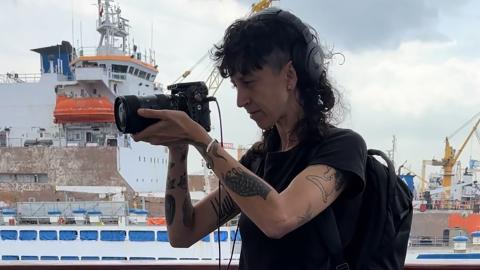 An image of Tan Safi holding a camera and filming aboard the deck of one of the ships of the Flotilla. Tan is wearing a black t-shirt, backpack and headphones. Other ships in port can be seen in the background.